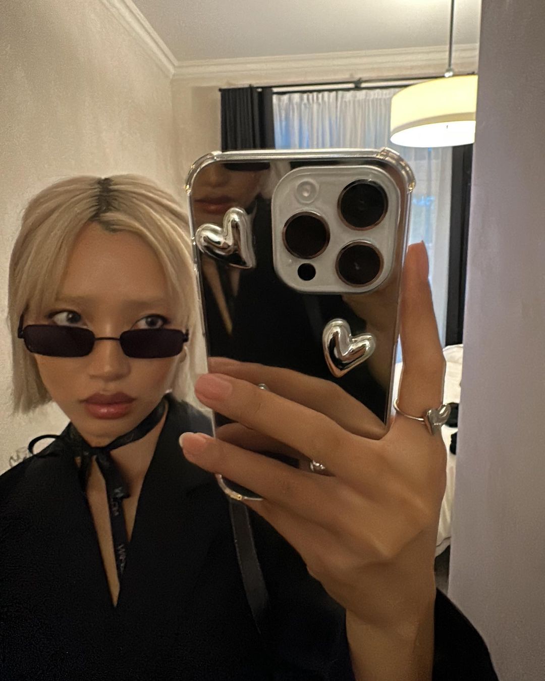 Blonde woman in black sunglasses and jacket takes a selfie in a mirror, showcasing a black cell phone with heart decorations.