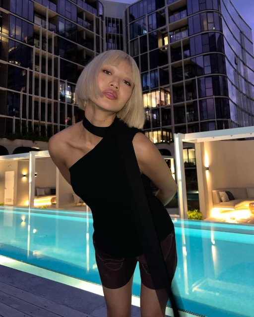 Blonde woman in black one-shoulder top and shorts poses next to an outdoor swimming pool with an apartment building in the background.
