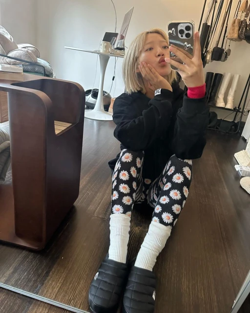 Woman takes a mirror selfie while sitting on the floor in a casual outfit with patterned leggings and black slip-on shoes in an indoor setting.