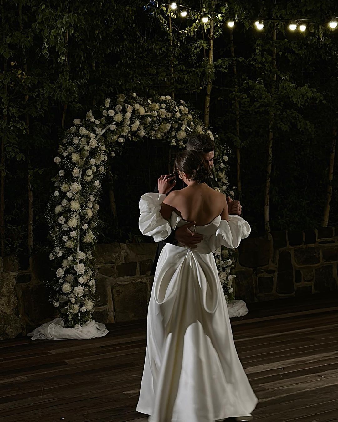 Couple in formal attire dancing under string lights near a floral archway in a dimly lit outdoor setting at nighttime.