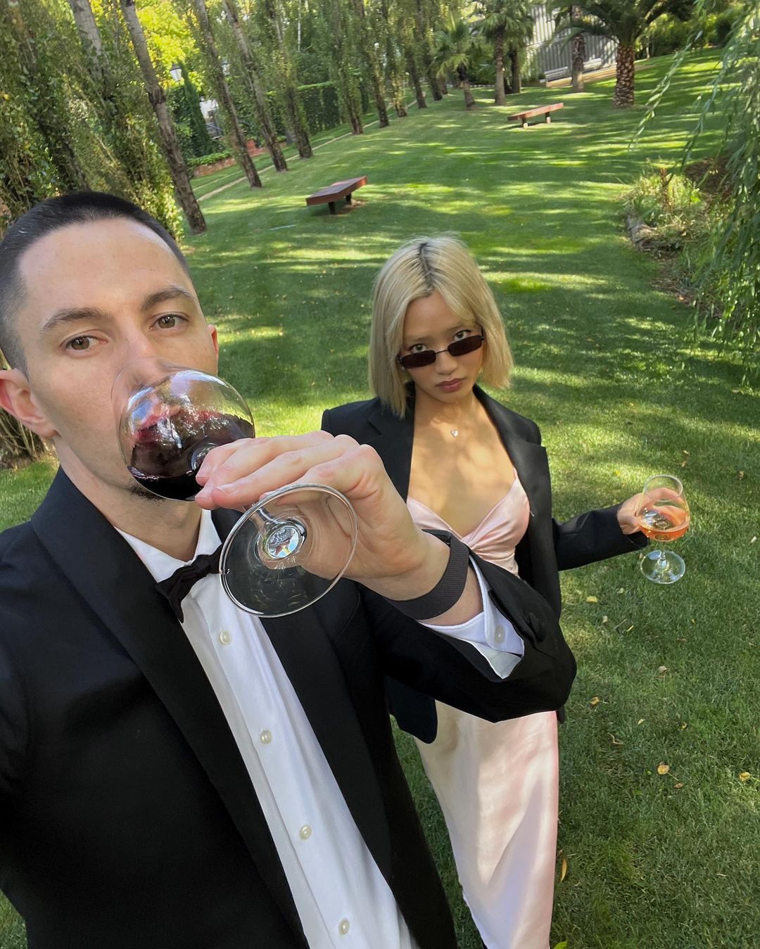 Two people, an adult man in a tuxedo and an adult woman in a dress and blazer, are in a grassy outdoor setting drinking wine.