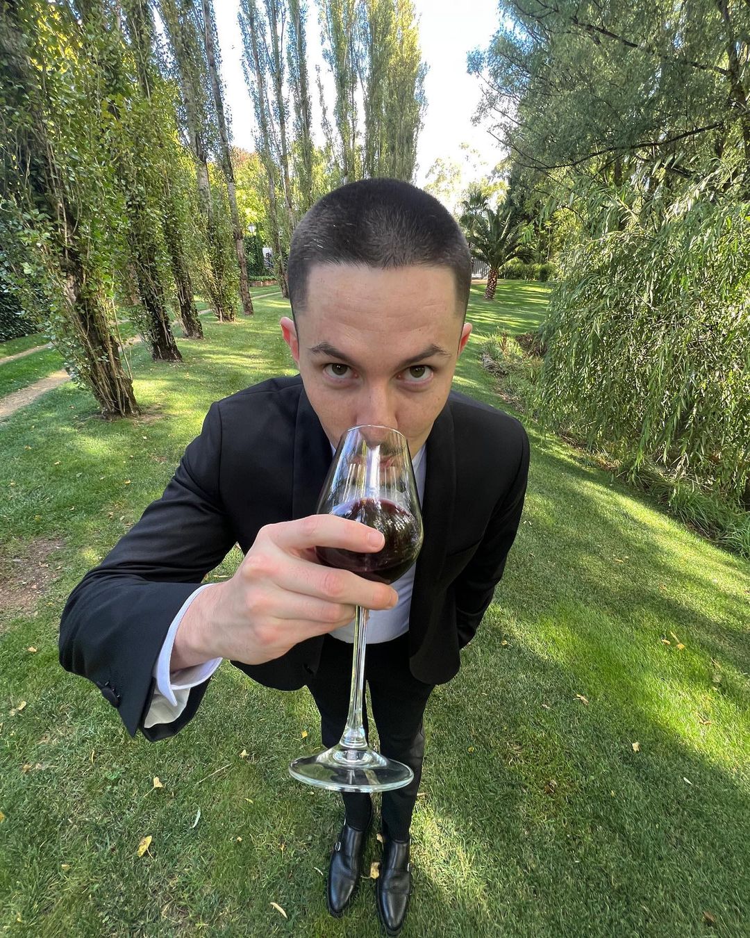 Adult male in a black suit drinks from a red wine glass on a sunny day outside with a background of green grass and tall trees.