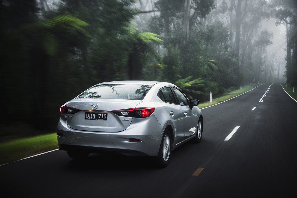 Silver Mazda sedan driving on a dark asphalt road surrounded by dense green forest and trees in a blurred motion effect.