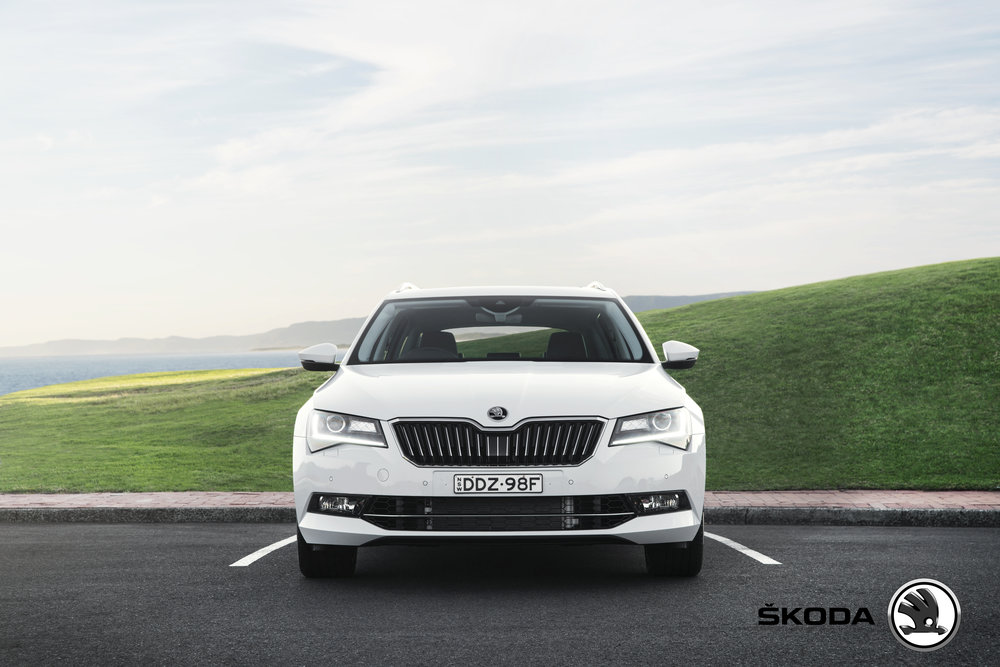 Front view of a white Skoda Superb parked on asphalt against a backdrop of a grassy hill and a distant ocean view under a bright sky.