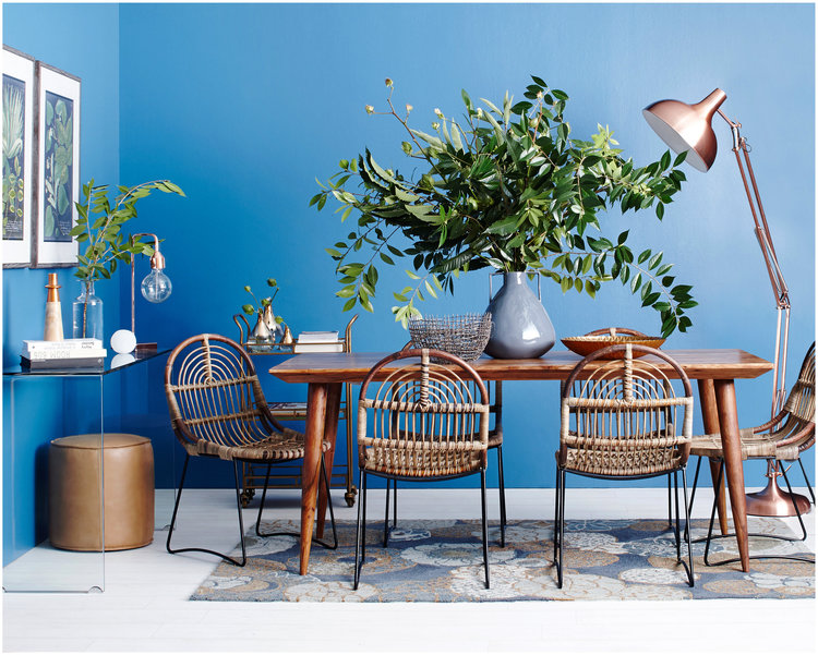 Dining room interior with a wood table, woven chairs, vase with greenery, copper-toned lamp, and blue wall in a home setting.