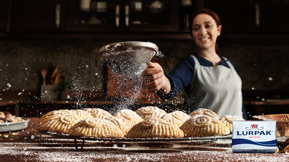 Woman dusting confectioner's sugar onto Maamoul cookies on a wire rack, with Lurpak butter in a warm kitchen setting, for food photography.