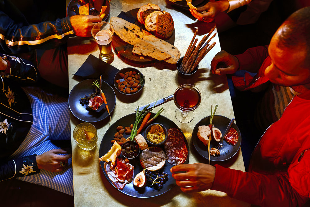 Overhead shot of several people seated at a table with drinks and appetizers on small plates and wooden platters, with food and drinks.