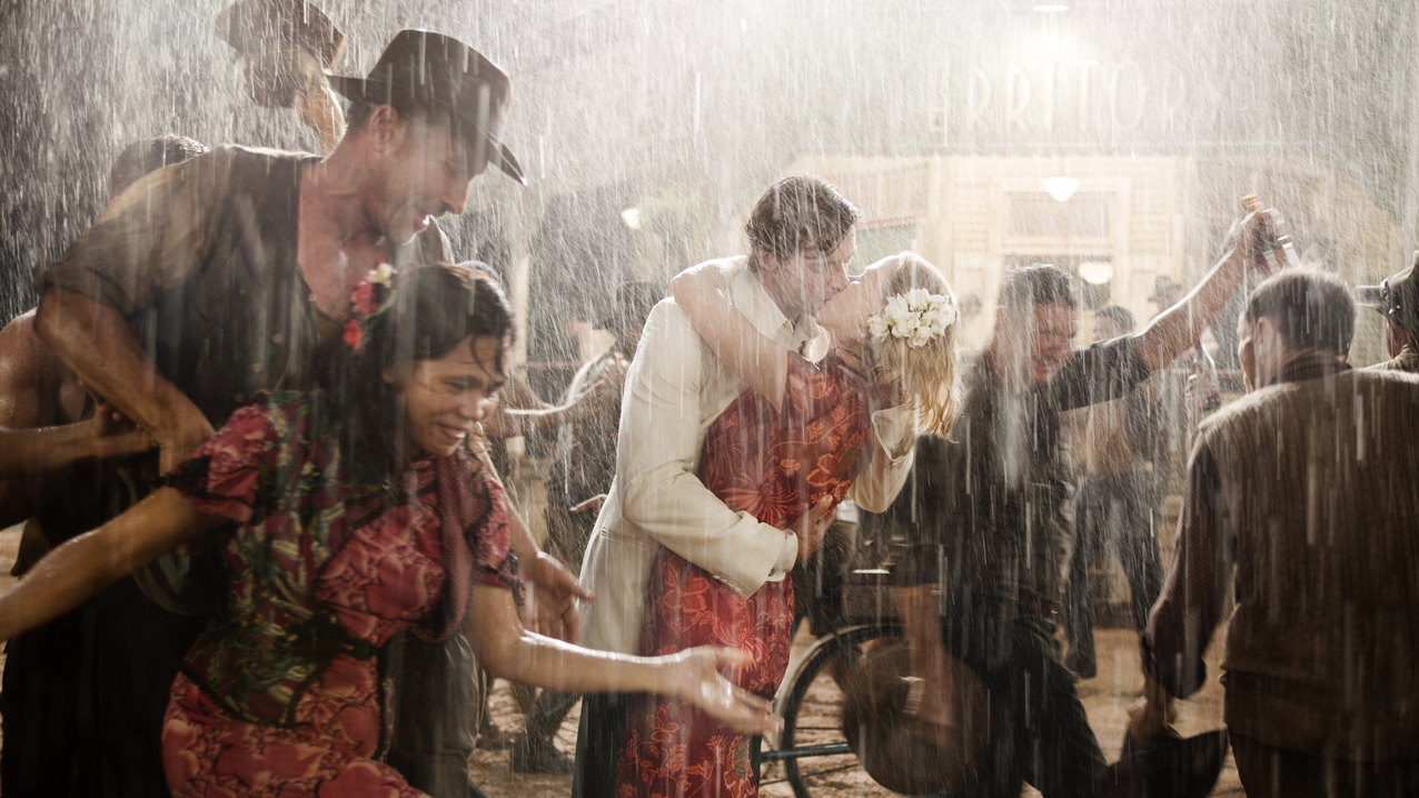 A couple embraces in the rain amidst a crowd of people, creating a candid, celebratory scene with vibrant colors and a sense of joy.