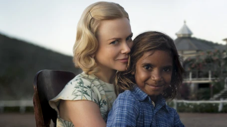 A blonde woman in a patterned dress embracing a young boy in a blue plaid shirt outdoors.