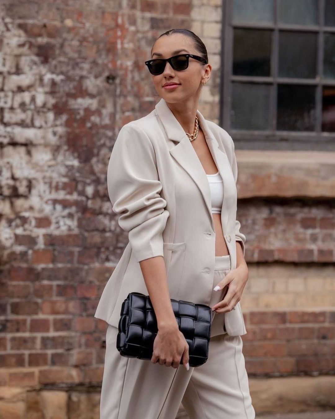 Woman in beige suit with black handbag stands against a brick wall, accessorized with sunglasses and gold jewelry, looking away.