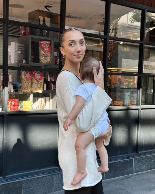 A woman is standing outside a building holding a baby wrapped in a blanket, with a window displaying food and books behind them.