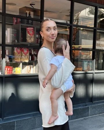 A woman is standing outside a building holding a baby wrapped in a blanket, with a window displaying food and books behind them.