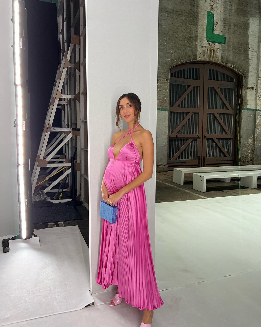A pregnant woman in a pink dress poses for a photo in an industrial space, holding a blue purse, with studio lighting on the side.