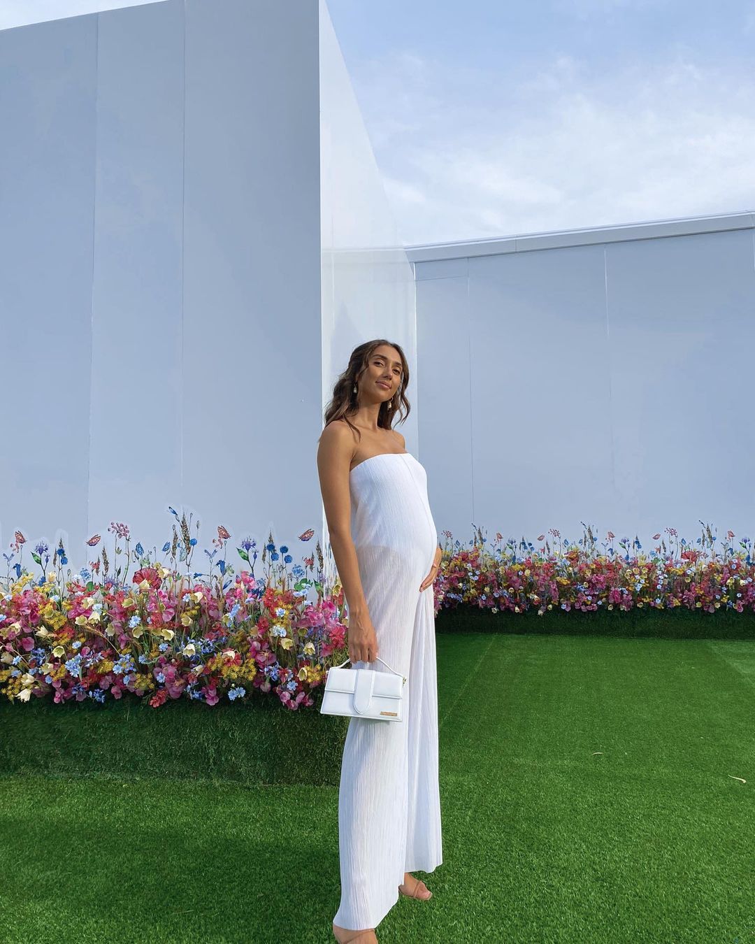 Brunette pregnant woman in white strapless jumpsuit and white purse stands outside on green grass near colorful flower garden.