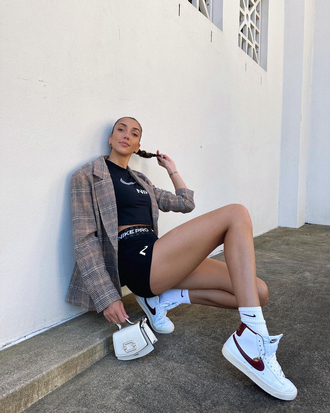 A woman poses in a blazer, crop top, and shorts sitting on a ledge in front of a white wall with white sneakers and a small handbag.
