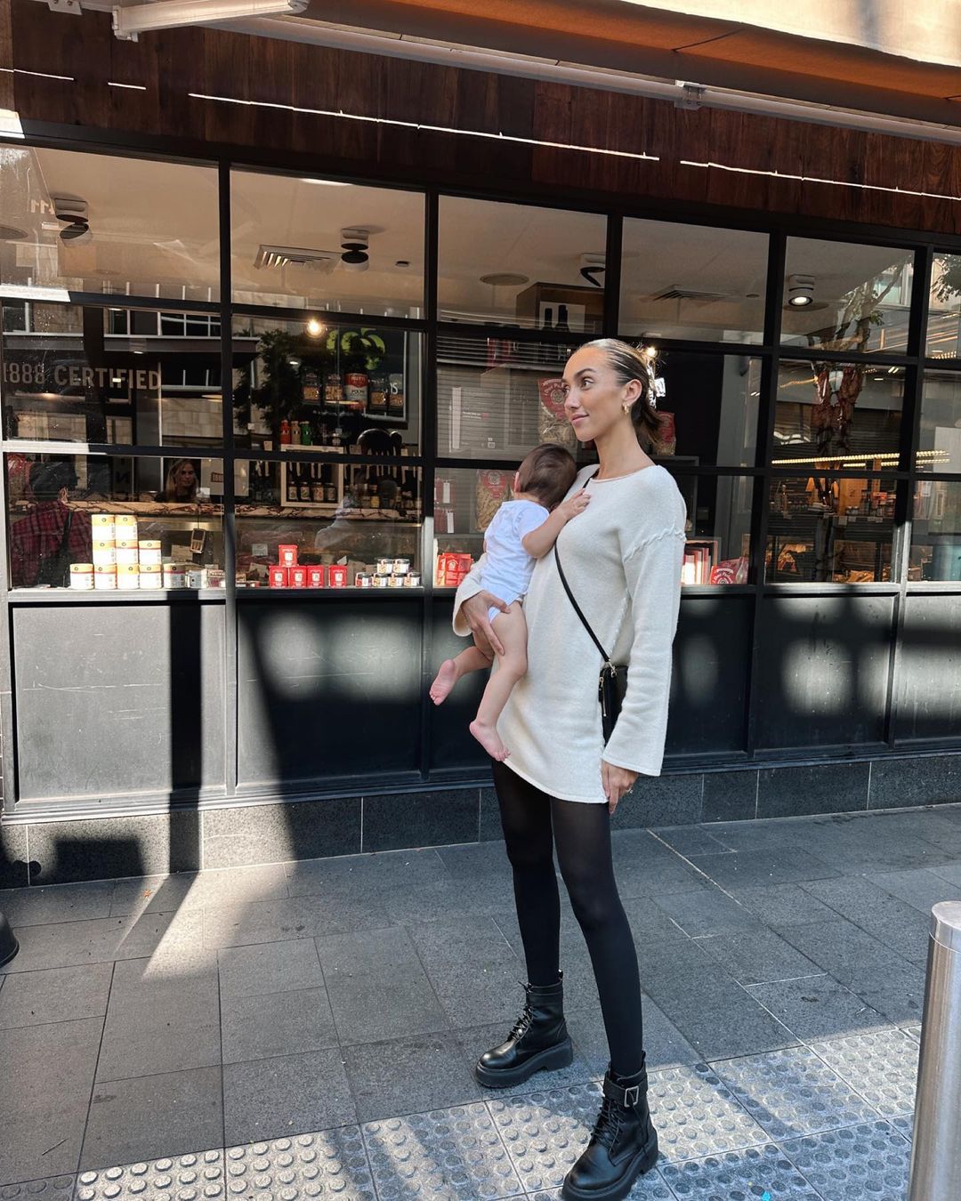 Adult female carrying a baby while wearing a cream mini-dress, black tights, and black platform boots on a city sidewalk.