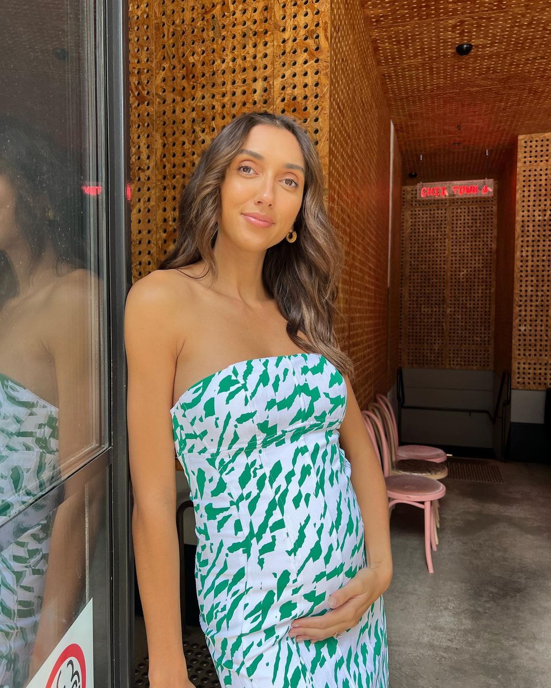 Brunette woman in a white and green patterned dress poses indoors near a dark door and golden textured wall.