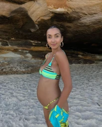 Pregnant woman in a striped bikini standing on a sandy beach in front of a textured rock formation