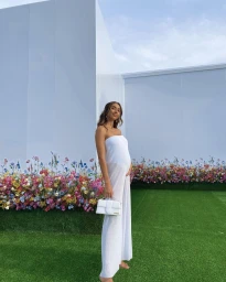 Pregnant woman in a white strapless jumpsuit holding a white handbag in front of floral displays and white walls.