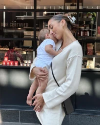Woman holding an infant child outside a storefront window, wearing a cream sweater and black crossbody bag.
