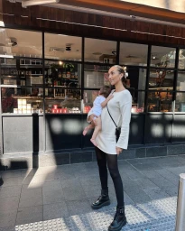 Woman in cream knit dress holding an infant on a city sidewalk outside storefront
