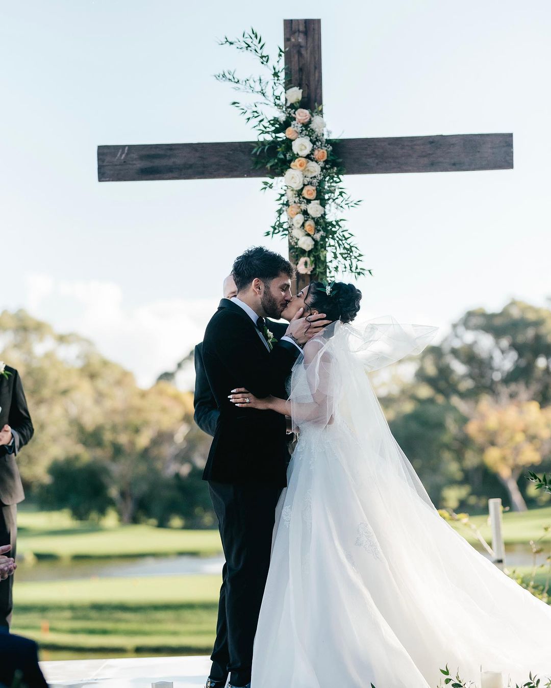 A couple kisses in front of a wooden cross decorated with white and peach roses at their outdoor wedding ceremony, on a sunny day.