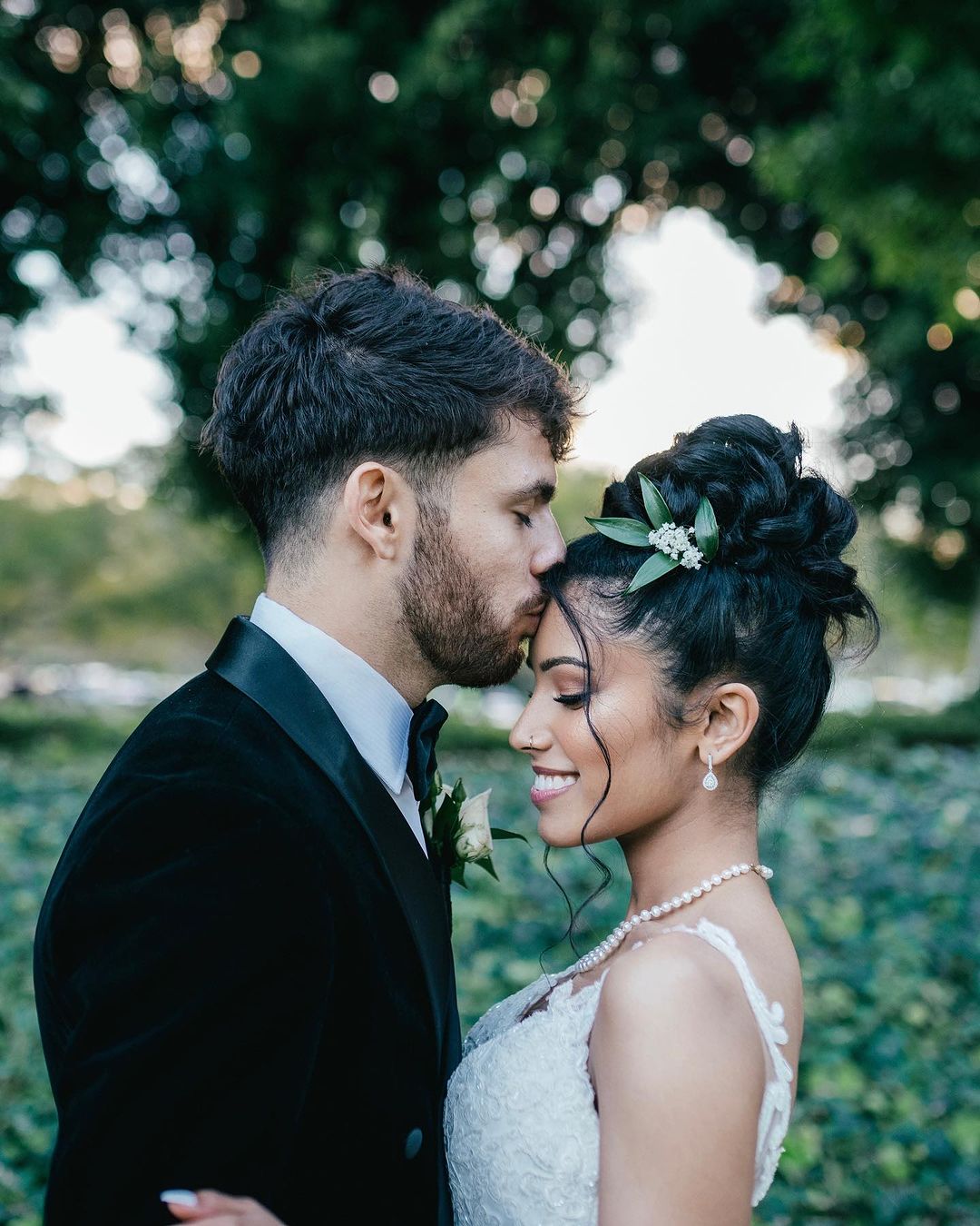 A couple shares a tender moment outdoors, with the groom kissing the bride's forehead amidst lush greenery and soft natural light.