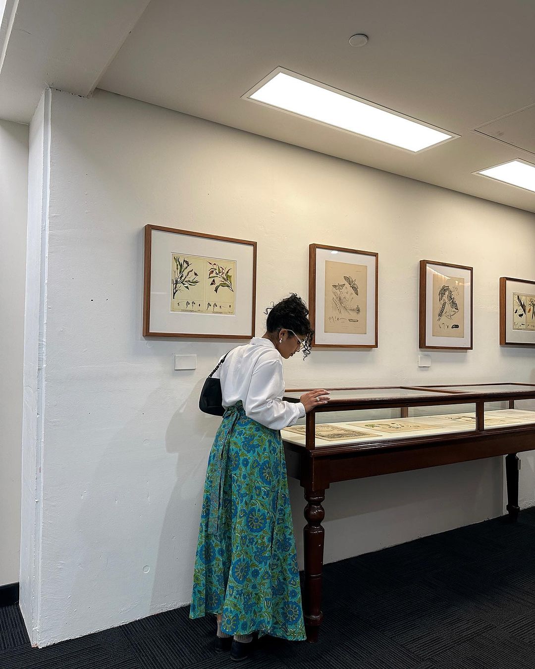 Woman with floral skirt views art behind glass display case in a gallery with white walls and recessed ceiling lights.