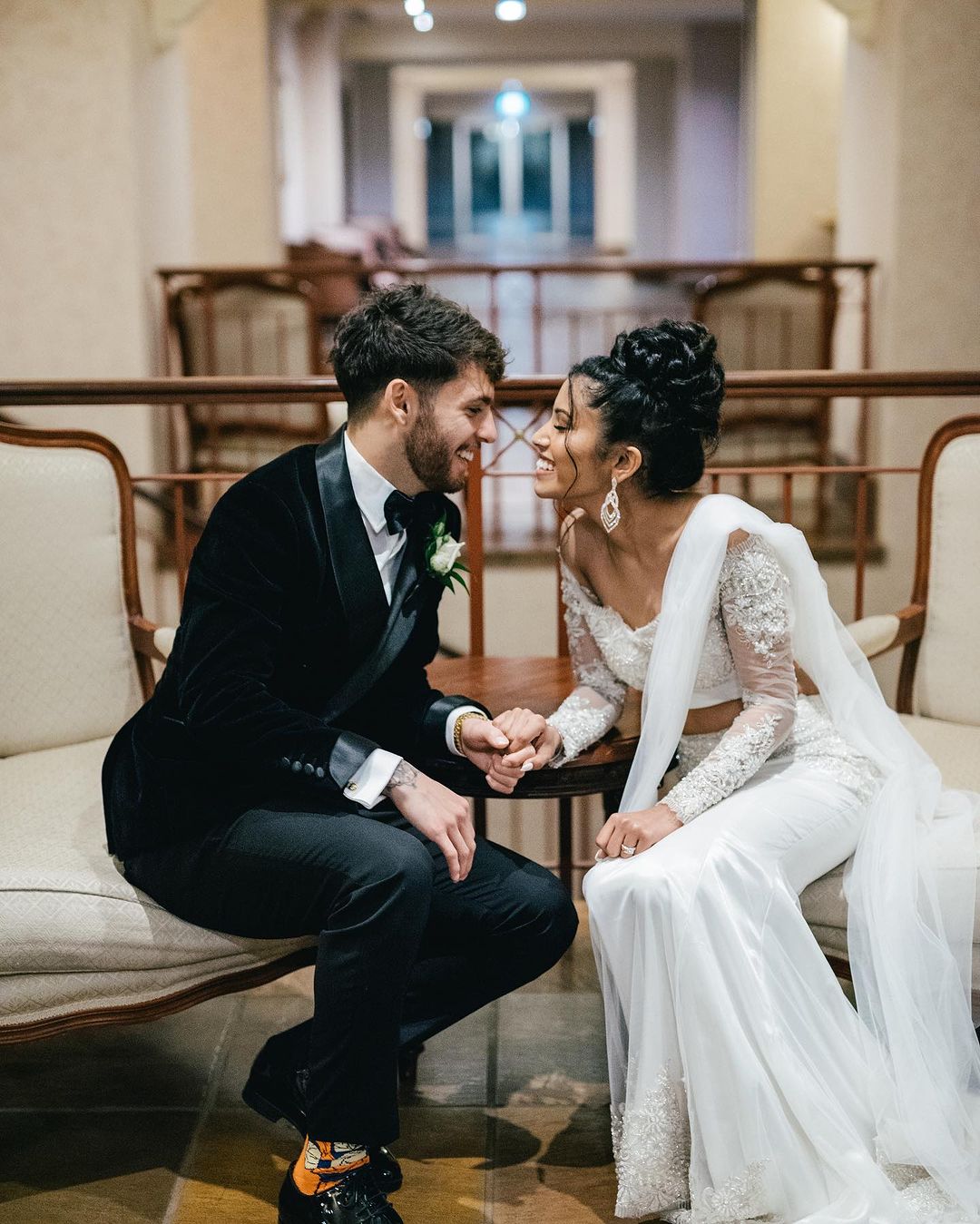 Wedding couple in formal attire share an intimate moment indoors, holding hands and smiling with soft, natural light and classic architecture.