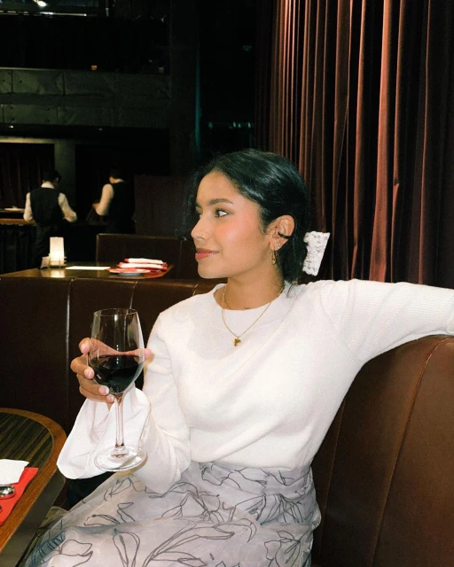 Elegant woman with dark hair in a white sweater and floral skirt sitting indoors, holding a glass of red wine in a dimly lit restaurant.