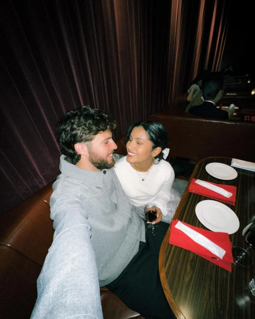 Indoor shot of a smiling couple at a restaurant, sitting at a wooden table with wine glasses and red napkins, with warm lighting.