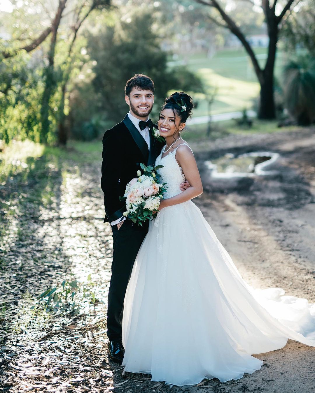 A smiling bride and groom, dressed in formal wedding attire, stand close together in a sunlit outdoor forest setting with a soft focus.