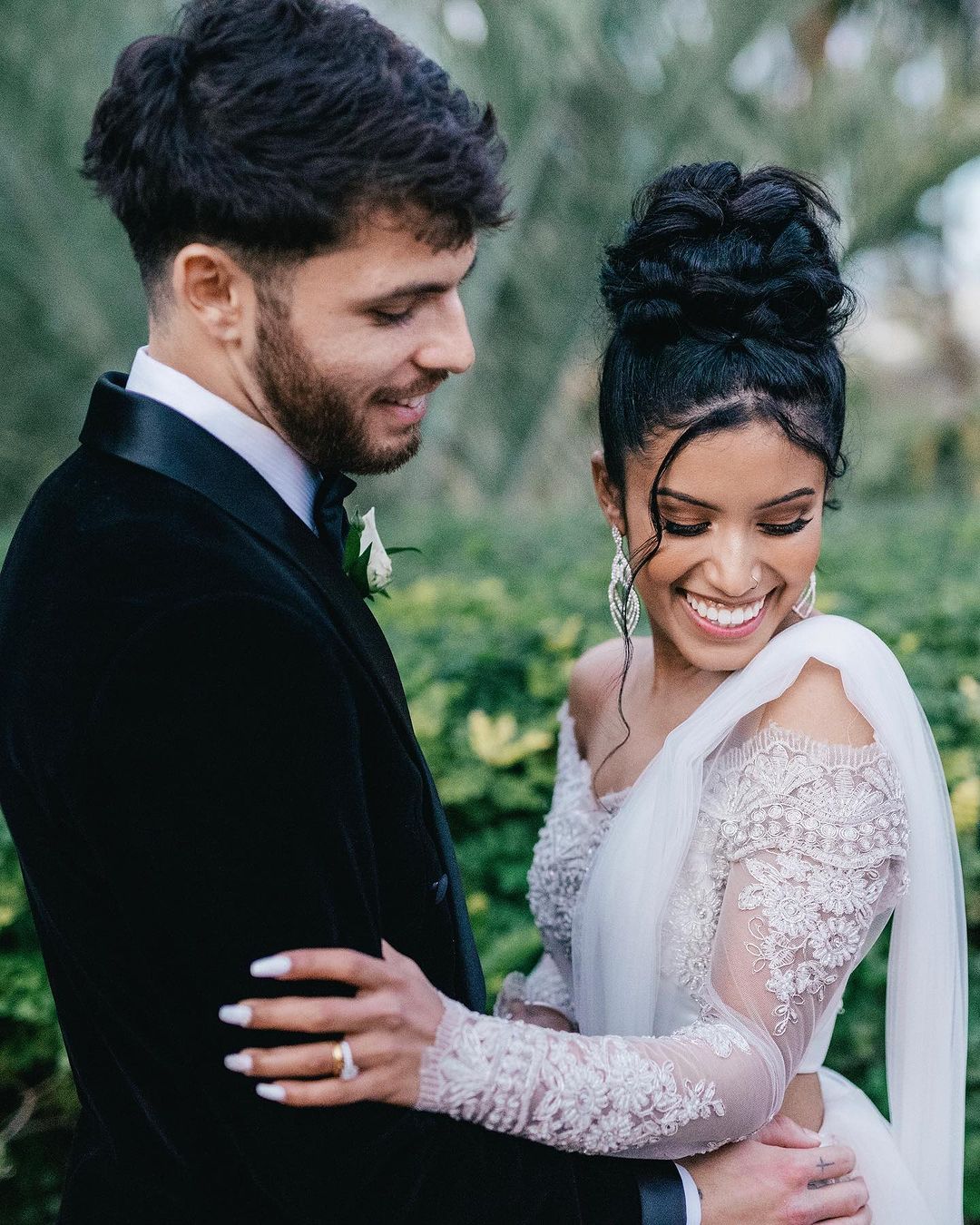 Portrait of a happy couple embracing outdoors, woman in ornate white gown, man in black tuxedo; greenery background.