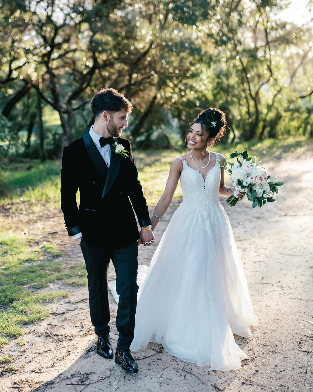 A smiling bride and groom hold hands on a tree-lined path. The bride carries flowers, and the groom wears a black tuxedo in natural light.