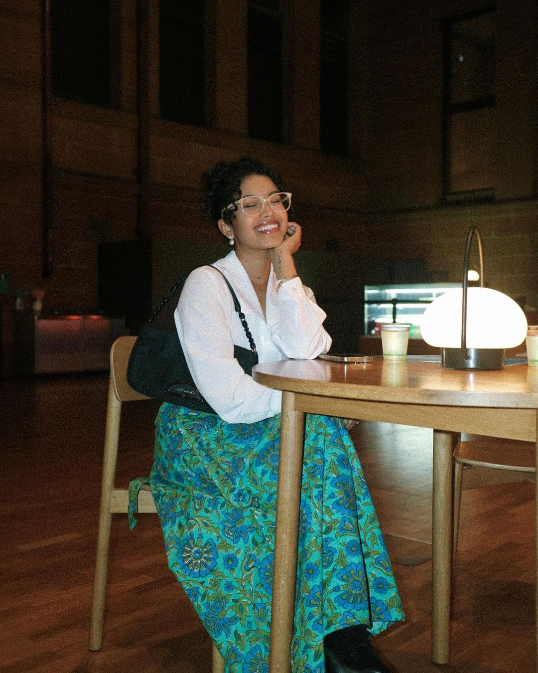 Smiling woman in white shirt sits at table with lamp and wood chairs in dimly lit room with patterned green skirt and black shoulder bag.