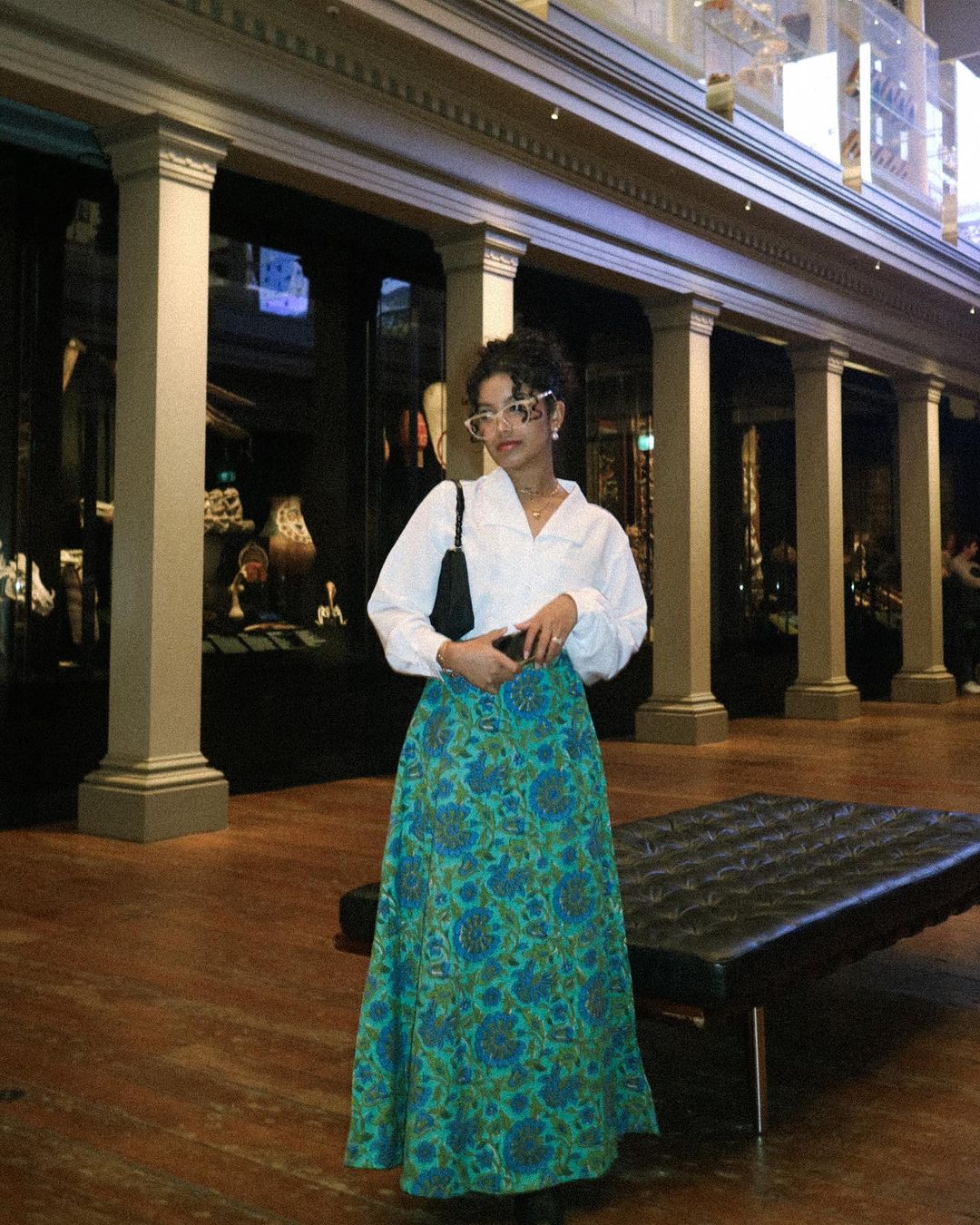 Adult female wearing a white blouse, long floral skirt and white glasses stands inside a museum hall with a black leather bag.