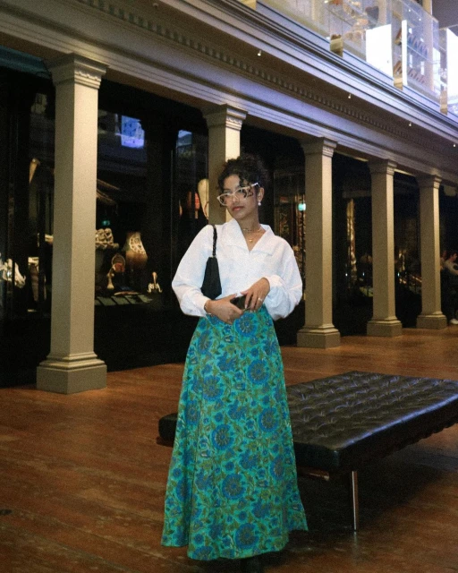 Adult female wearing a white blouse, long floral skirt and white glasses stands inside a museum hall with a black leather bag.