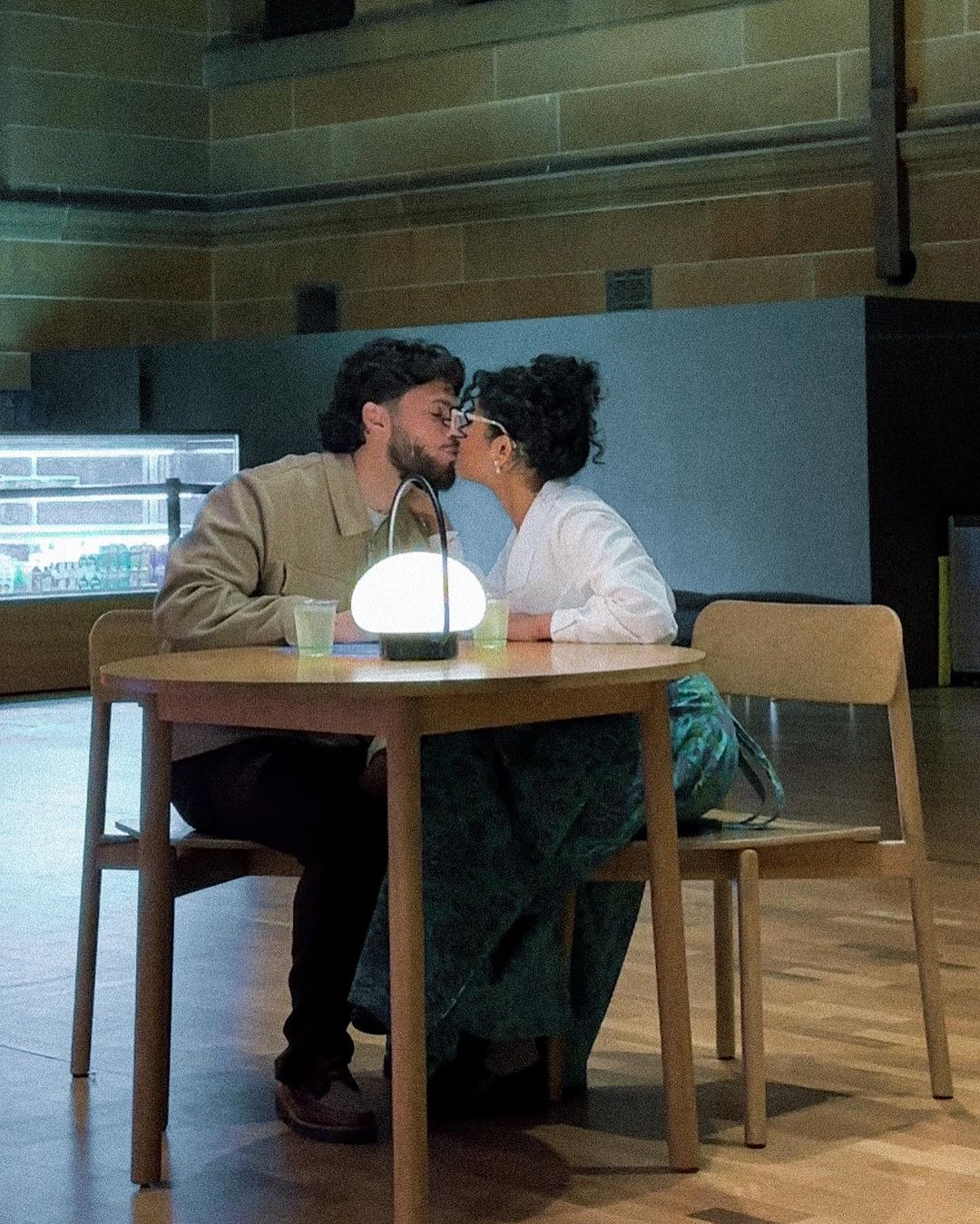 A kissing couple sits at a table indoors, lit by a glowing lamp. The man and woman are stylishly dressed in a warm and inviting atmosphere.