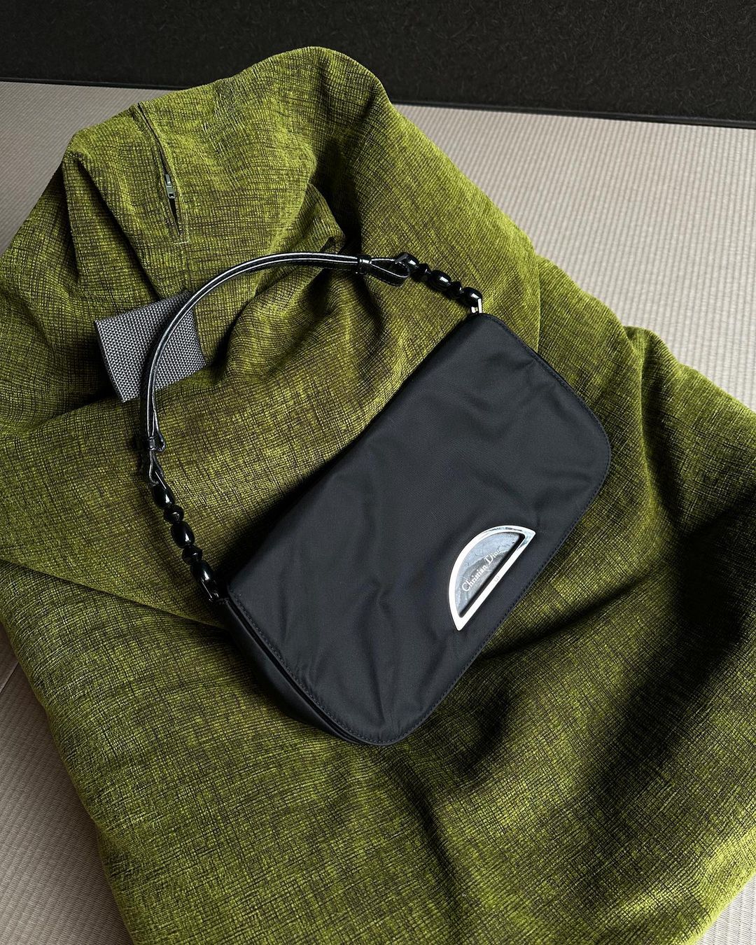 Black purse with a silver logo and beaded strap placed on top of a textured, green blanket and beige background. Fashion.