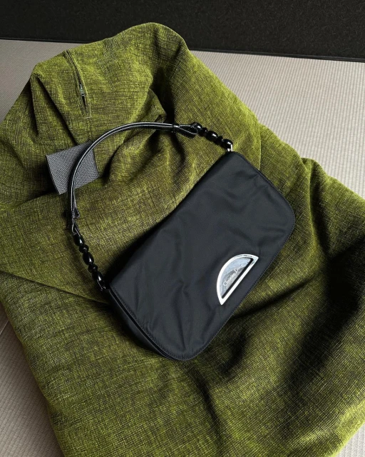 Black purse with a silver logo and beaded strap placed on top of a textured, green blanket and beige background. Fashion.