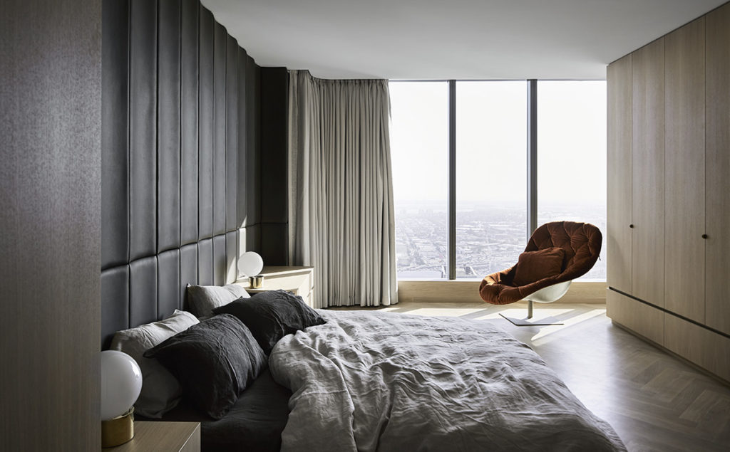 Interior view of a modern bedroom with a large bed, headboard, bedside tables with lamps, a designer chair, and a city view through large windows.