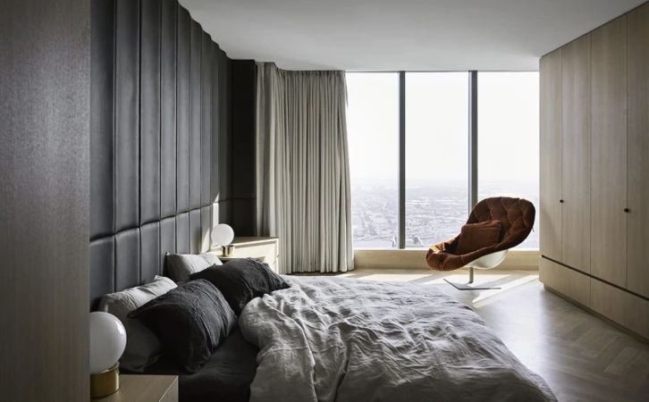 Interior view of a modern bedroom with a large bed, headboard, bedside tables with lamps, a designer chair, and a city view through large windows.