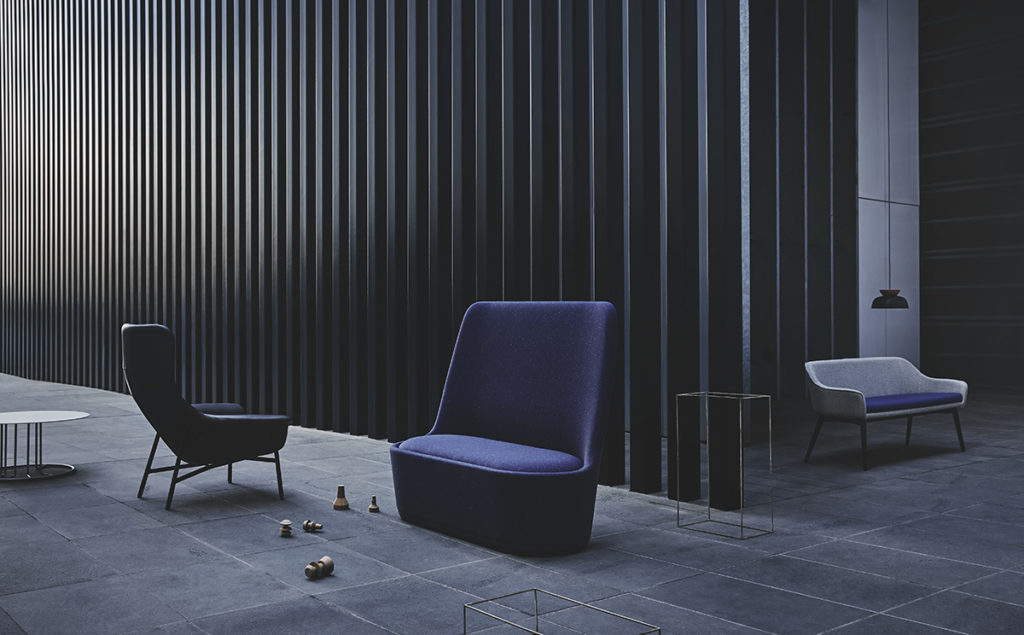 Indoor lounge area with a blue armchair, blue two-seater, and black chair on a grey-blue tile floor against a black slat wall.