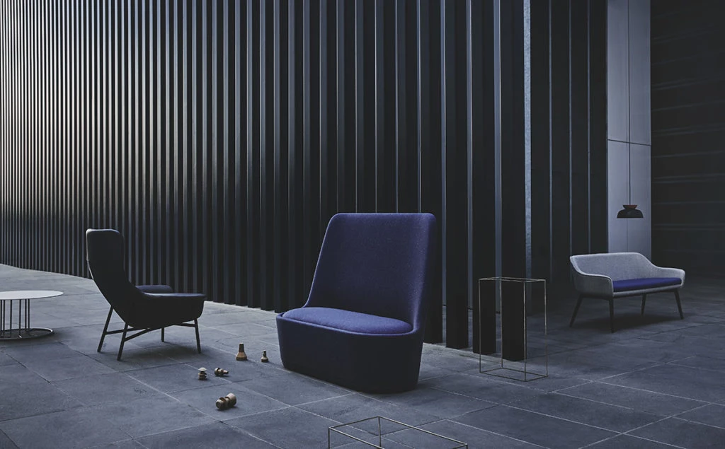 Indoor lounge area with a blue armchair, blue two-seater, and black chair on a grey-blue tile floor against a black slat wall.