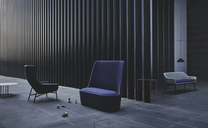 Indoor lounge area with a blue armchair, blue two-seater, and black chair on a grey-blue tile floor against a black slat wall.