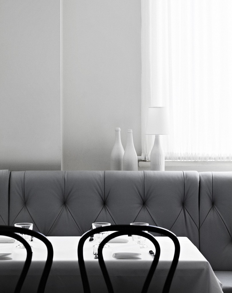 Formal dining room with set table in grayscale. White tablecloth, tableware, chairs, couch, decorative bottles, and window with curtain.