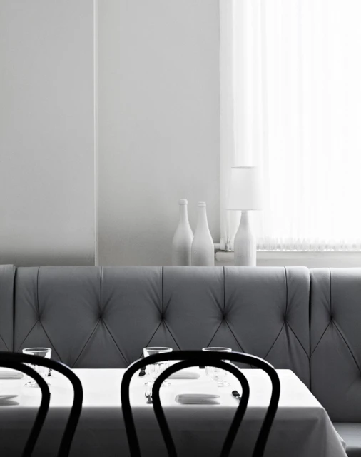 Formal dining room with set table in grayscale. White tablecloth, tableware, chairs, couch, decorative bottles, and window with curtain.