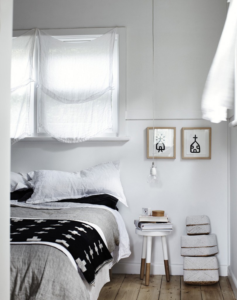 Cozy bedroom interior with a bed featuring black and white patterned throw blanket, white walls, light wood floors, and framed artwork.
