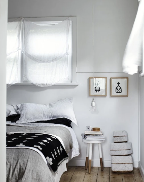 Cozy bedroom interior with a bed featuring black and white patterned throw blanket, white walls, light wood floors, and framed artwork.