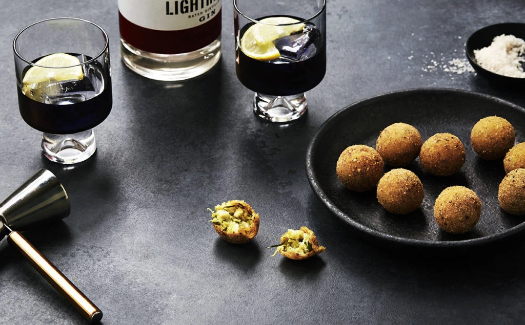 Still life shot of two cocktails in stemmed glasses with lemon wedges, an open arancini, a plate of arancini, and gin on a dark surface.